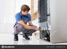 Mężczyzna w kombinezonie roboczym, w kucki, sprawdza dolną część chłodziarki; widoczne rurki, sprężarka i podłączenia elektryczne, jasne płytki na podłodze.