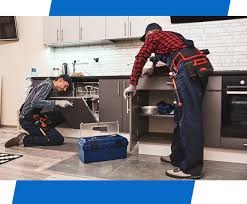 Dwóch mężczyzn w roboczych ubraniach naprawia pralkę w kuchni; jeden z nich klęczy przed urządzeniem, drugi stoi obok otwartej szafki z narzędziami, widoczna niebieska skrzynka narzędziowa.