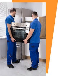 Dwóch mężczyzn w niebieskich uniformach transportuje piekarnik na tle lodówki ze stali nierdzewnej.