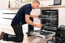 Mężczyzna w trakcie naprawy zmywarki, klęczy przed otwartym urządzeniem, wyciągając szufladę, widoczne narzędzia przy pasie, logo Electrolux na spodniach.