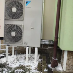 Zewnętrzna jednostka pompy ciepła Daikin Altherma z dwoma wentylatorami, zamontowana na białym stelażu przy elewacji budynku w zimowej scenerii, z widocznym brązowym rurociągiem spustowym.