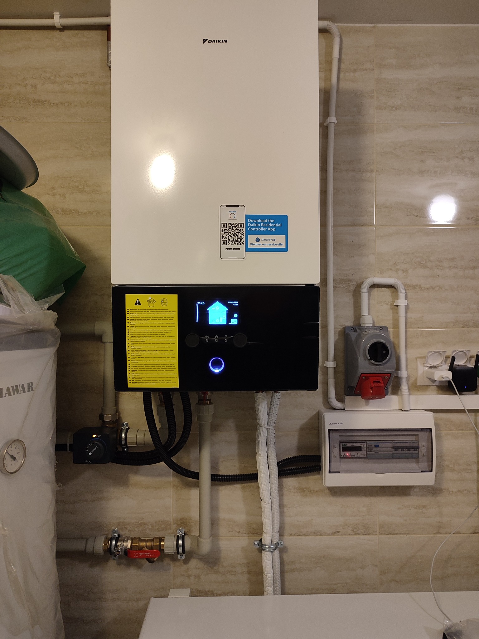 Zamontowany na ścianie, biały piec grzewczy marki Daikin z podłączonymi rurami, widoczny również sterownik z niebieskim podświetleniem, tablica elektryczna i osprzęt. Na piecu naklejka z kodem QR...