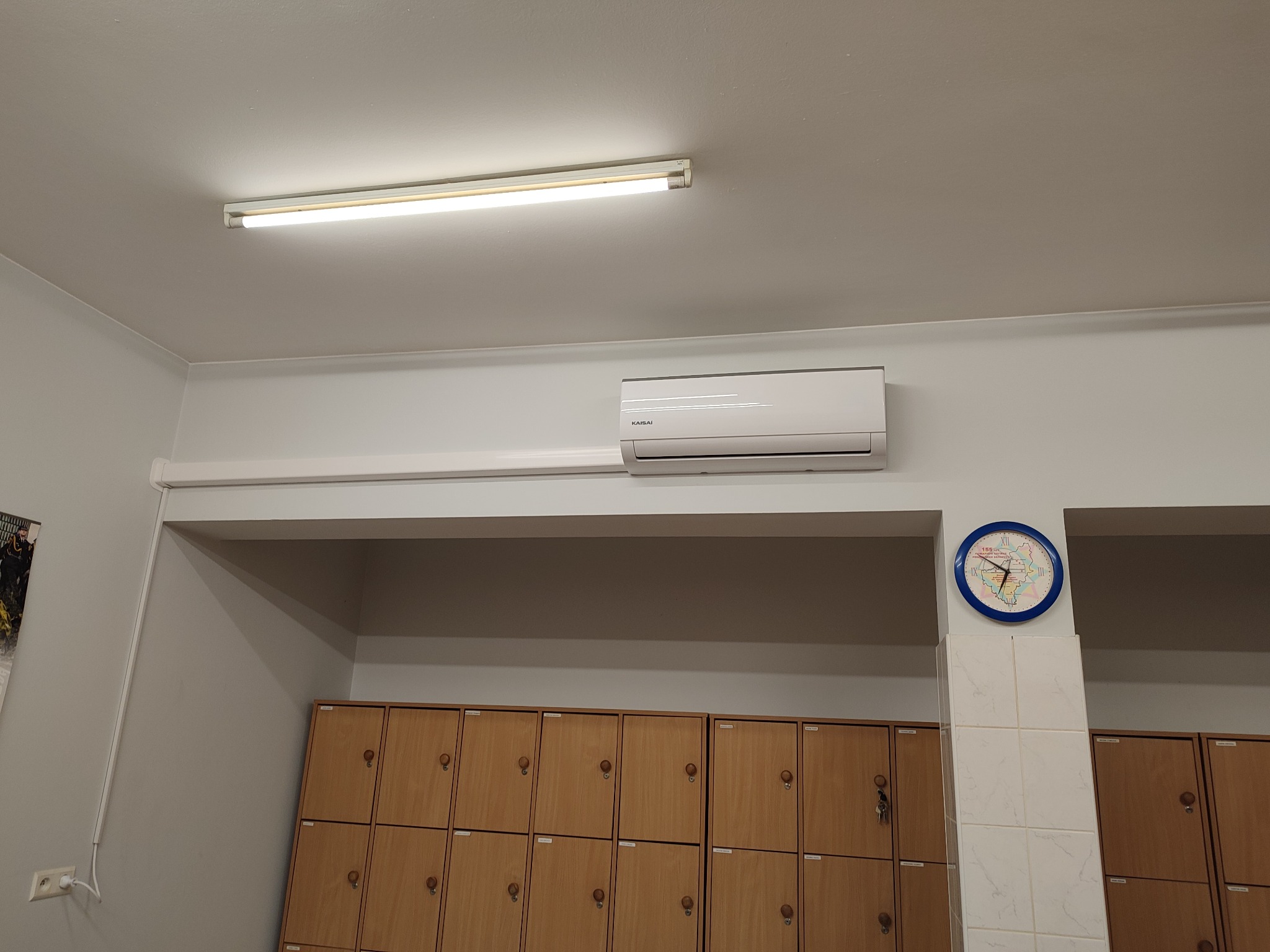 Biały klimatyzator marki KAISAI zamontowany na ścianie nad rzędem brązowych szafek pracowniczych, widoczny również zegar ścienny z motywem mapy Polski.