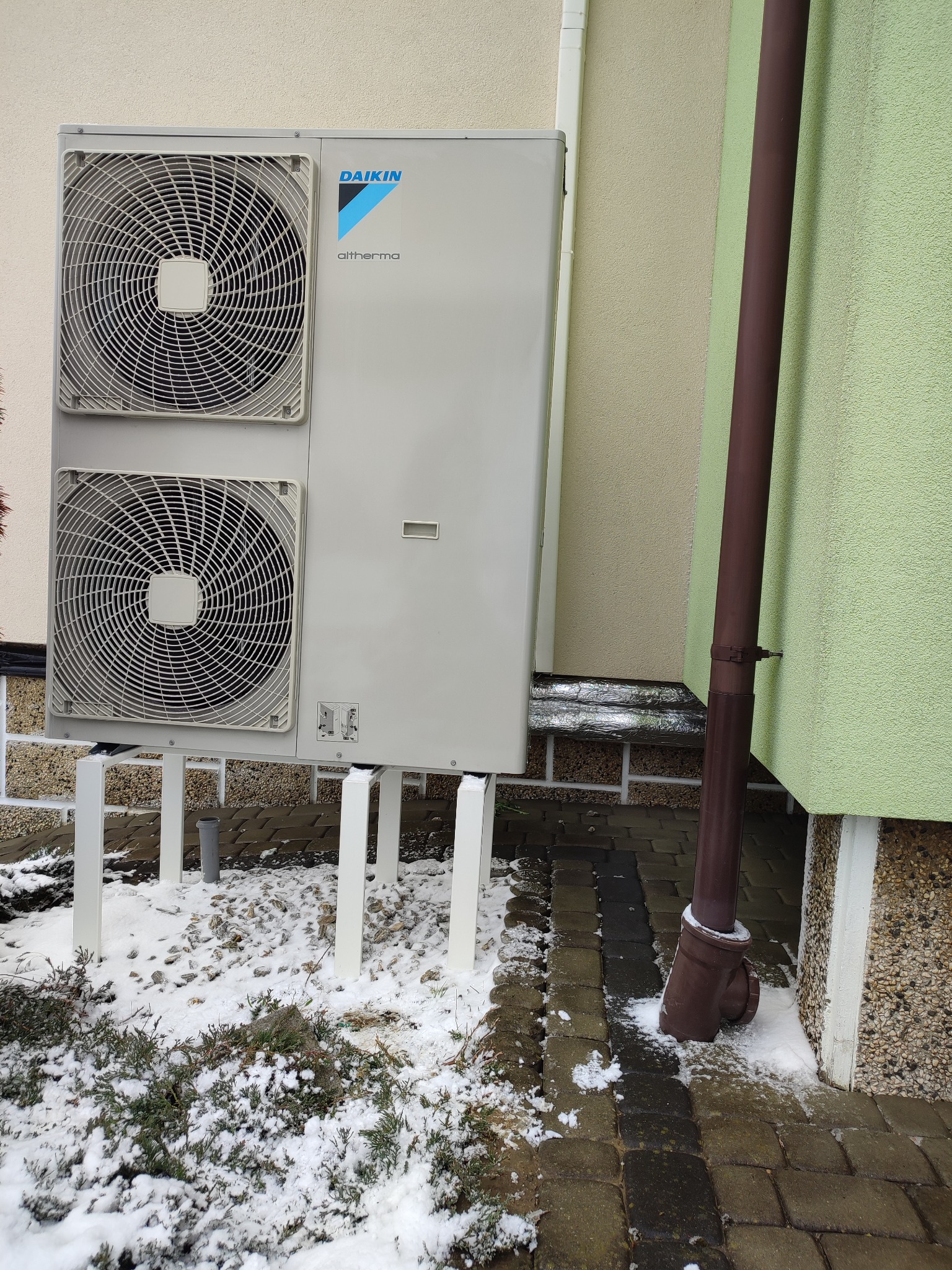 Zewnętrzna jednostka pompy ciepła Daikin Altherma z dwoma wentylatorami, zamontowana na białym stelażu przy elewacji budynku w zimowej scenerii, z widocznym brązowym rurociągiem spustowym.