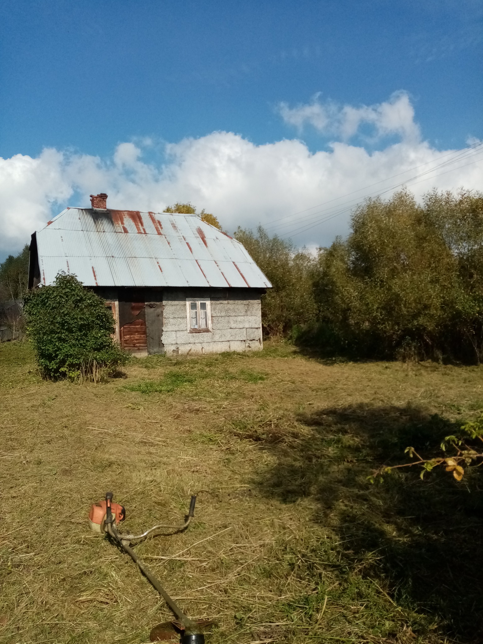 Nisko skoszona trawa wokół starego, zaniedbanego domu z blachy falistej i ceglanym kominem, z widoczną pomarańczową podkaszarką spalinową na pierwszym planie, w słoneczny dzień.