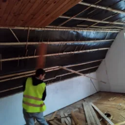 Poddasze w trakcie ocieplania: widoczna czarna folia izolacyjna przymocowana do drewnianych belek, pracownik w żółtej kamizelce odwrócony tyłem, deski na podłodze, okno w tle.