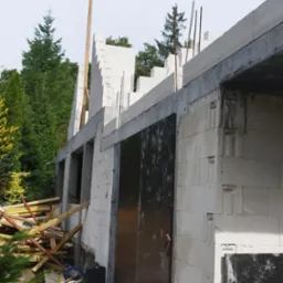 Surowy stan budynku z białych bloczków, widoczne stalowe pręty zbrojeniowe wystające z góry, czarne płyty zabezpieczające, stos drewna i materiałów budowlanych w tle, zielona roślinność.