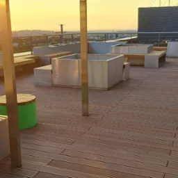 Drewniany taras na dachu budynku z metalowymi ławkami, donicami i zielonym stołkiem, oświetlony zachodzącym słońcem.