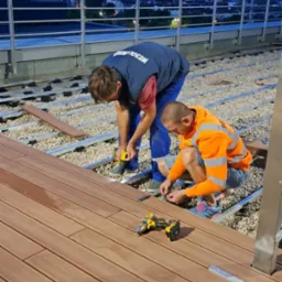 Montaż kompozytowej deski tarasowej na dachu budynku, dwaj pracownicy przykręcają deski do metalowej konstrukcji na tle wieczornego miasta, widoczne narzędzia: wiertarka, miarka, ołówek i wiadro.