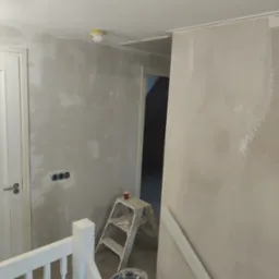 Ściana w trakcie szpachlowania z widocznymi śladami po nałożonym gipsie, drabina malarska, wiadro z zaprawą, zabezpieczone gniazdka elektryczne i fragment białej balustrady.