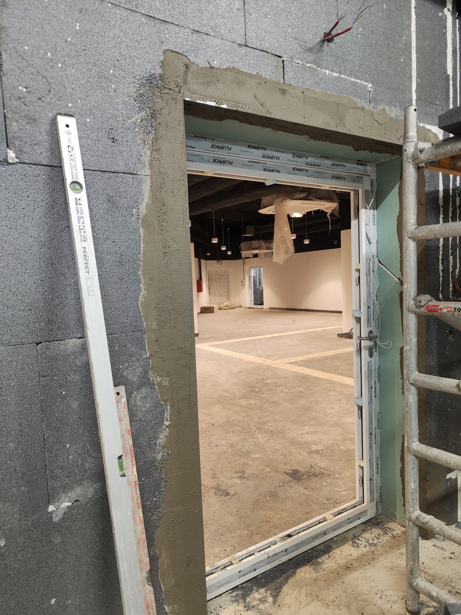 Montaż ościeżnicy w ścianie z ociepleniem, widoczny poziomica, surowy tynk i fragment rusztowania w pomieszczeniu komercyjnym.