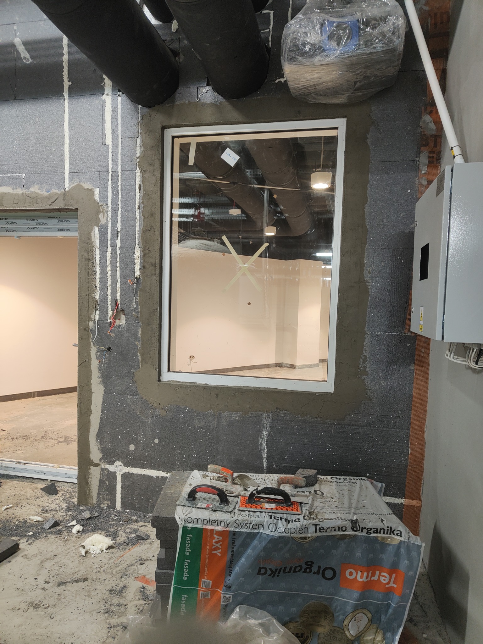 Montaż okna w surowym pomieszczeniu, ściany ocieplone grafitowym styropianem, widoczne narzędzia i worki z materiałem ociepleniowym Termo Organika, instalacja elektryczna w puszce.