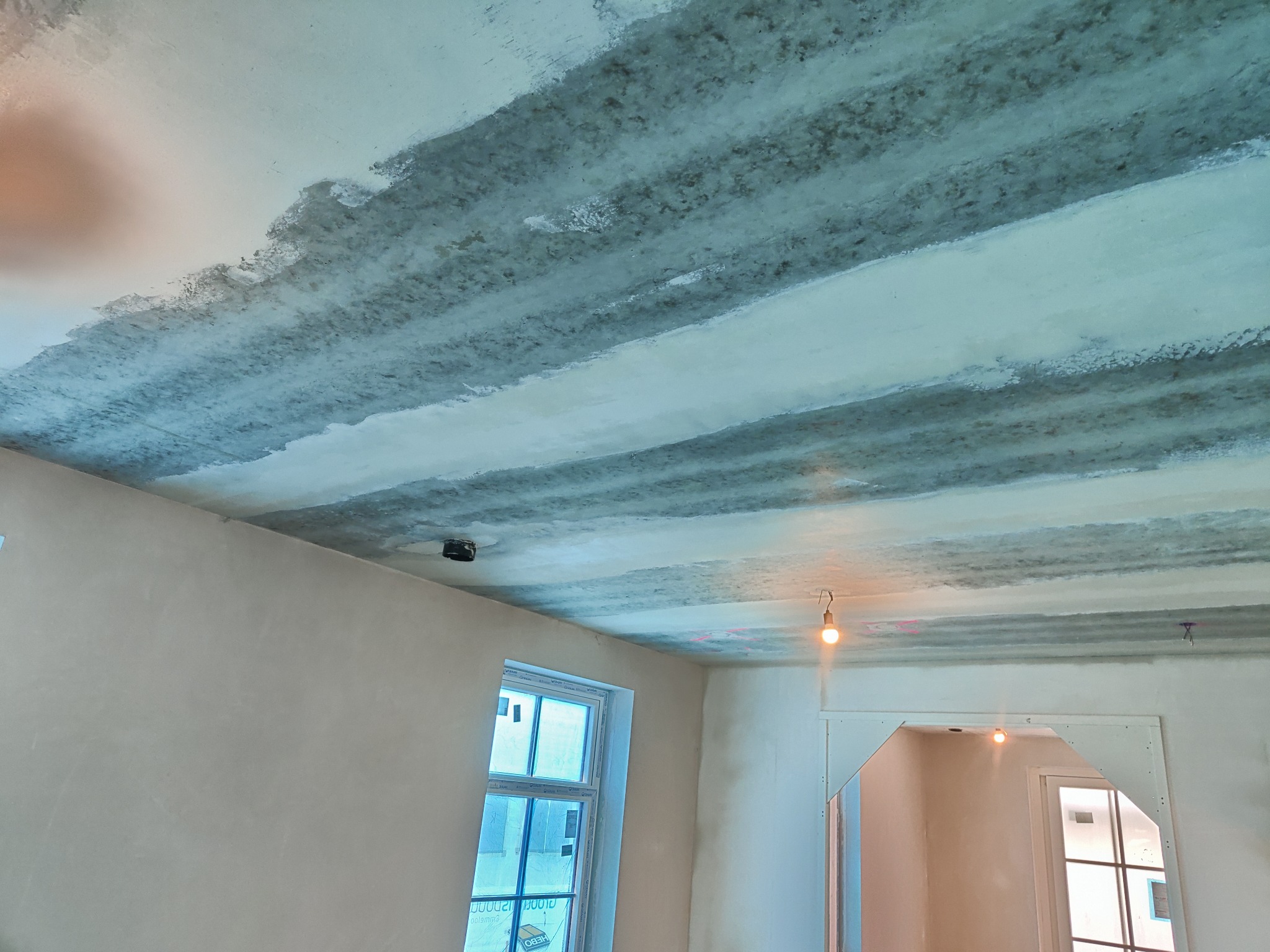 Niedokończony sufit z widocznymi pasami szpachli, ściany przygotowane do malowania, surowy stan wykończenia wnętrza z widocznym oknem i prowizorycznym oświetleniem.