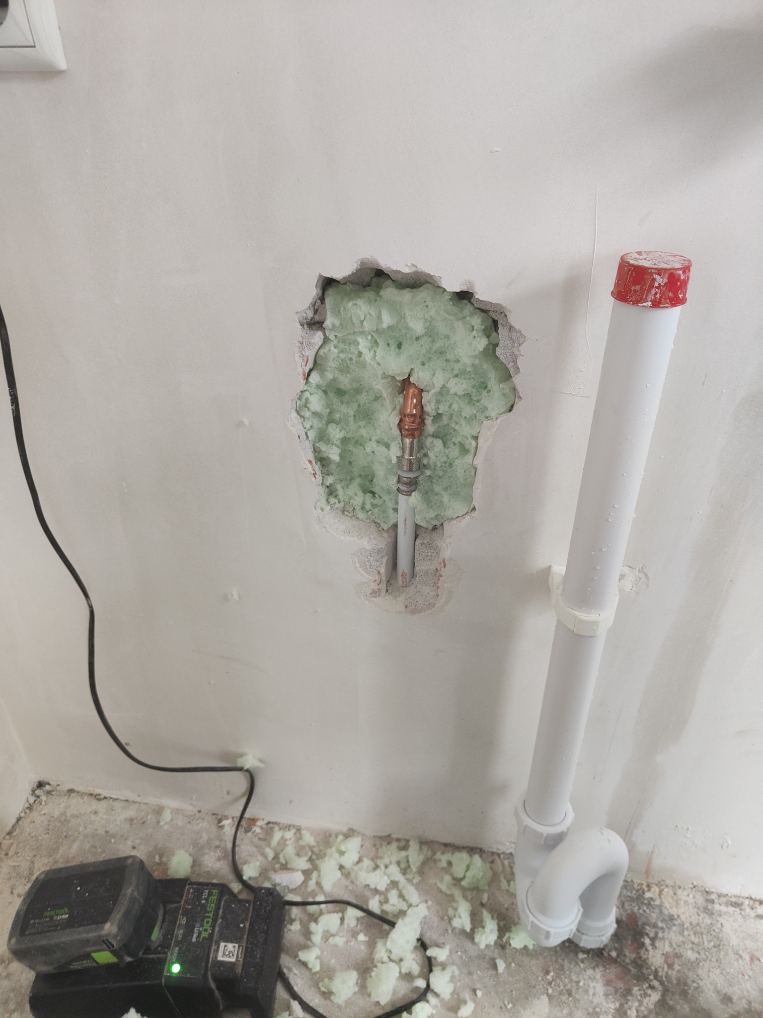 Odsłonięty fragment ściany z rurą wodną otoczony zieloną pianką izolacyjną, obok biała rura odpływowa i poziomica laserowa Festool na podłodze.