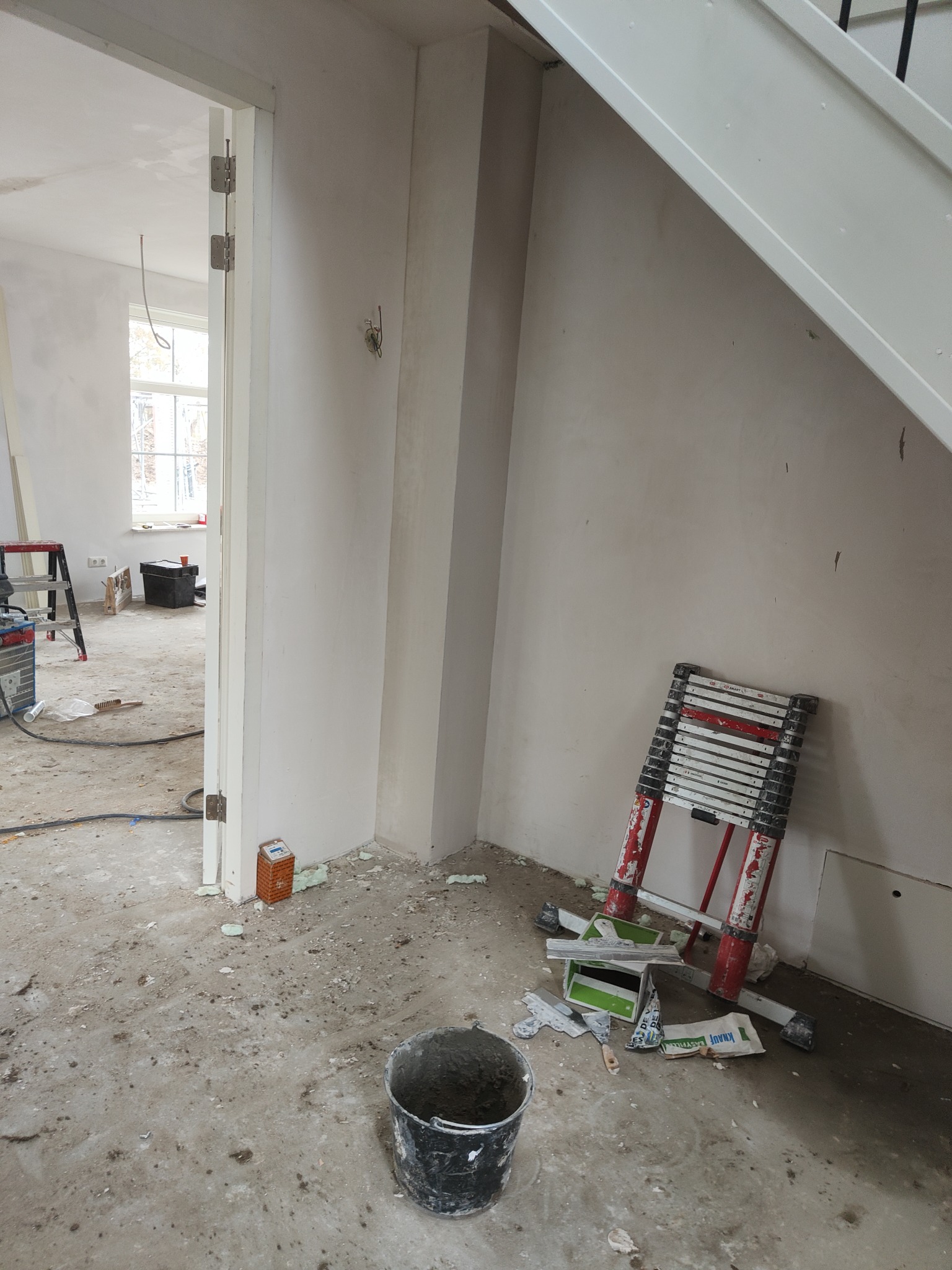 Wnętrze w trakcie prac wykończeniowych: ściany po szpachlowaniu, widoczny kubeł z zaprawą, drabina teleskopowa oparta o ścianę, rozsypane materiały budowlane, otwarte drzwi do kolejnego pomieszczenia.
