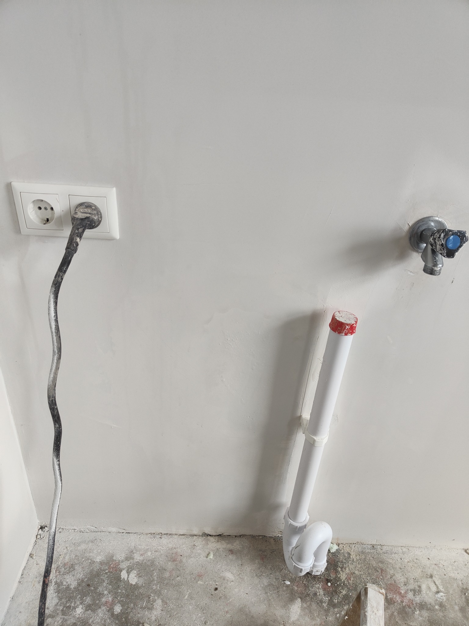 Świeżo wyszpachlowana ściana z gniazdkiem elektrycznym z wpiętym kablem w farbie, zaworem wodnym i rurą kanalizacyjną z czerwonym korkiem, widoczne ślady prac remontowych na podłodze.