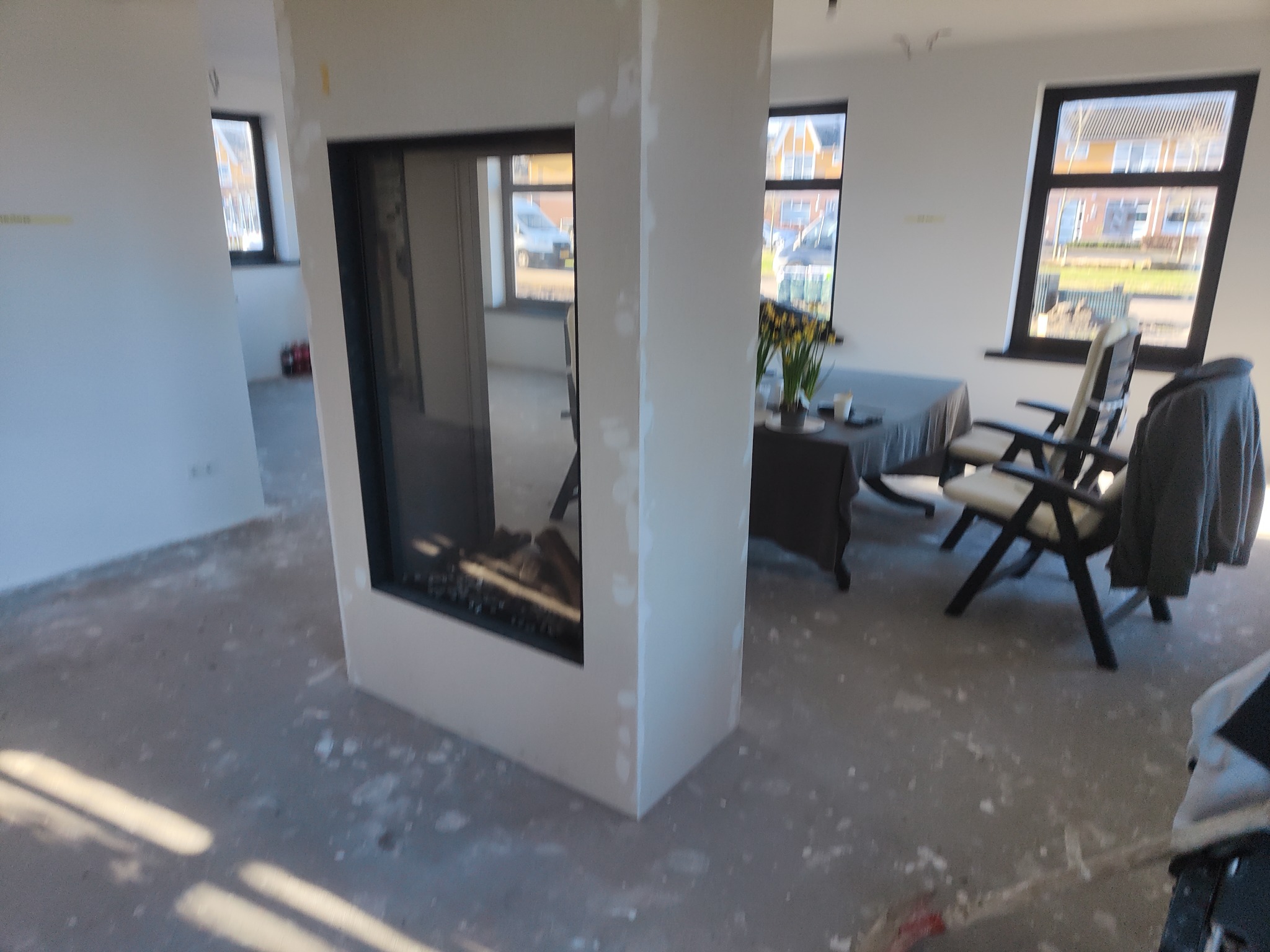 Wnętrze w trakcie prac wykończeniowych: widoczny wkład kominkowy w obudowie z płyt gipsowo-kartonowych, szpachlowane ściany, podłoga z widocznymi zabrudzeniami po pracach, w tle stół z krzesłami.