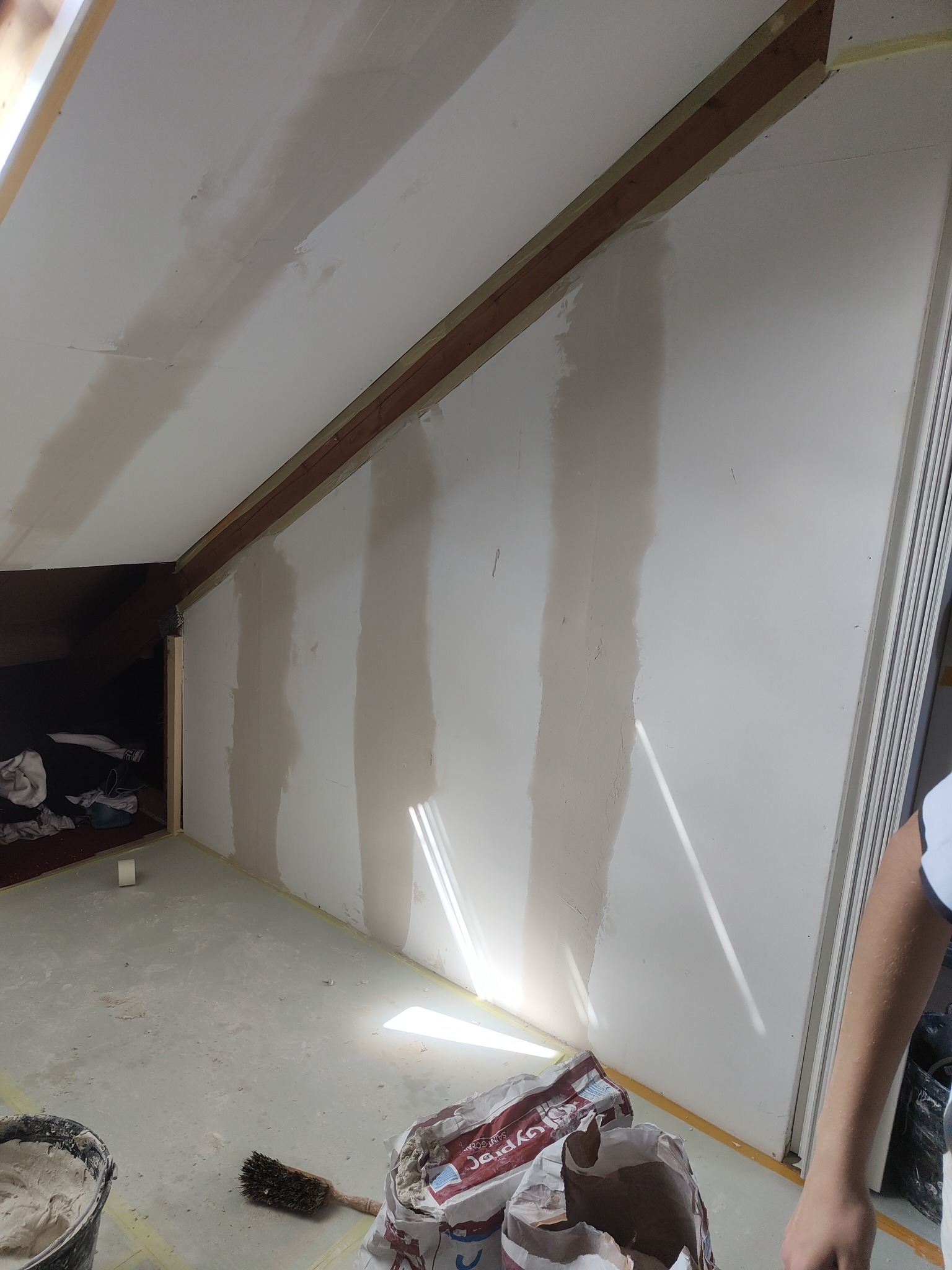 Poddasze w trakcie prac wykończeniowych: ściany częściowo pokryte masą szpachlową, widoczne worki z gipsem, wiadro z zaprawą i pędzel na podłodze zabezpieczonej taśmą malarską.