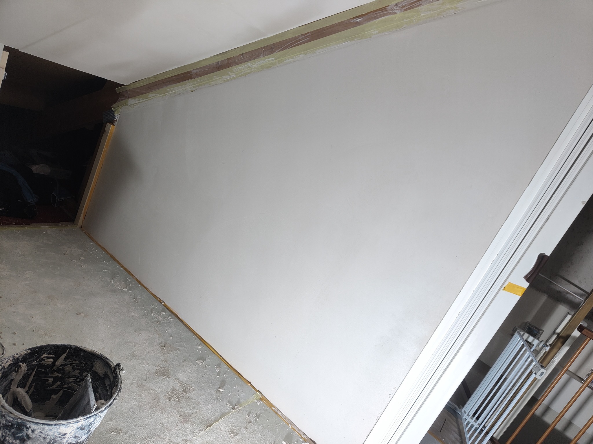 Świeżo wyszpachlowana ściana w trakcie remontu, zabezpieczona taśmą malarską przy suficie, z widocznym wiadrem i szpachelką na podłodze.