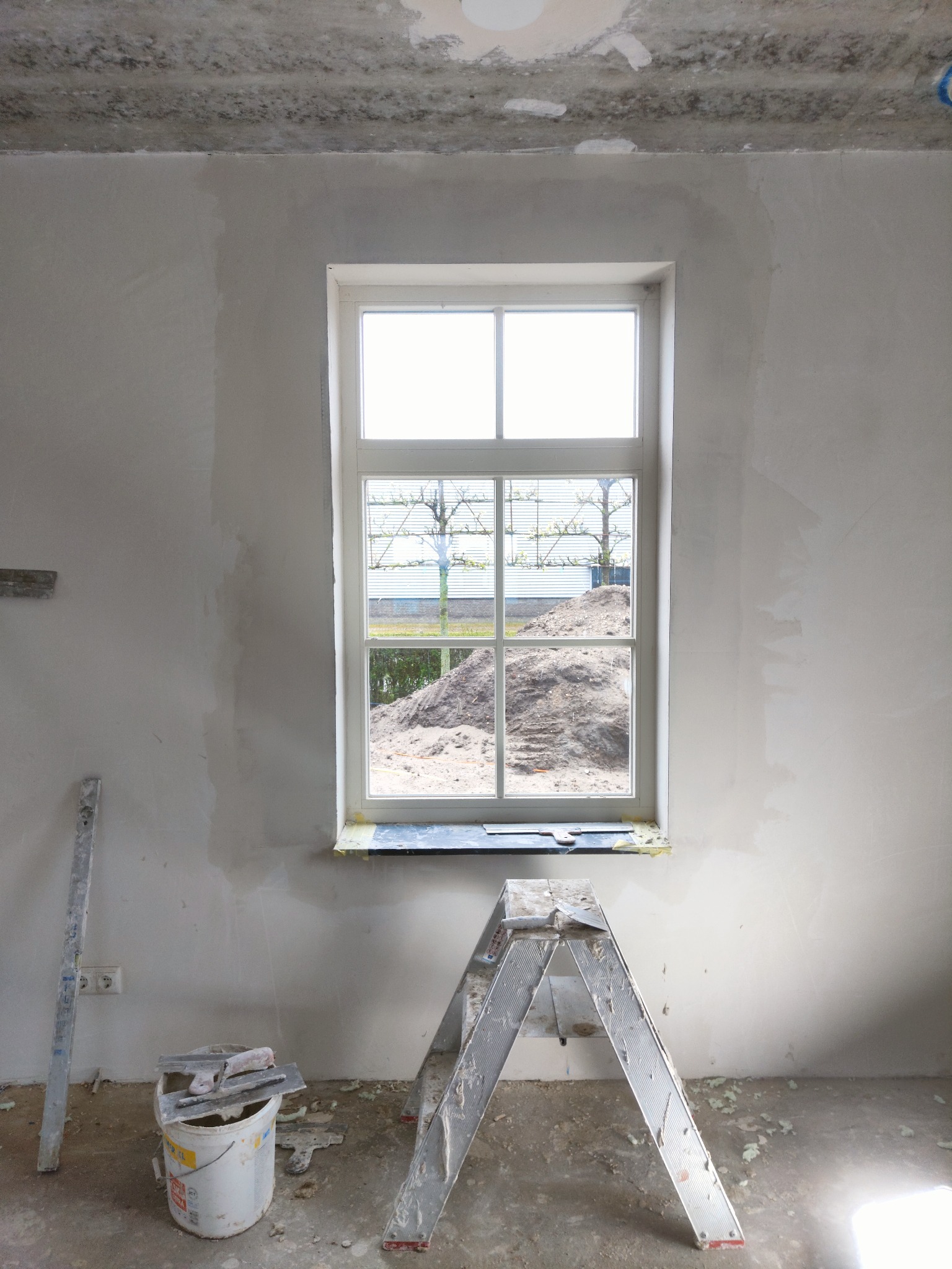 Wnętrze w trakcie prac wykończeniowych: ściany po szpachlowaniu, widoczna drabina, wiadro z narzędziami i okno z widokiem na hałdę ziemi.