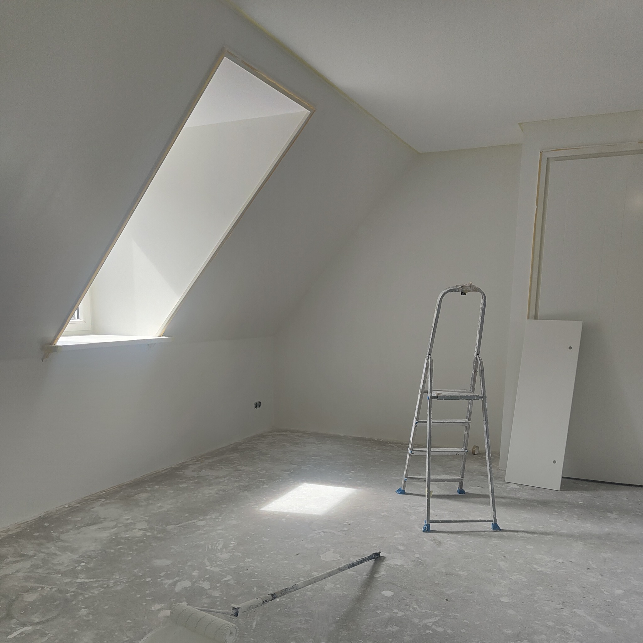 Pokój na poddaszu po świeżym pomalowaniu na biało, widoczna drabina aluminiowa, wałek do malowania na podłodze, okno dachowe wpuszczające światło.