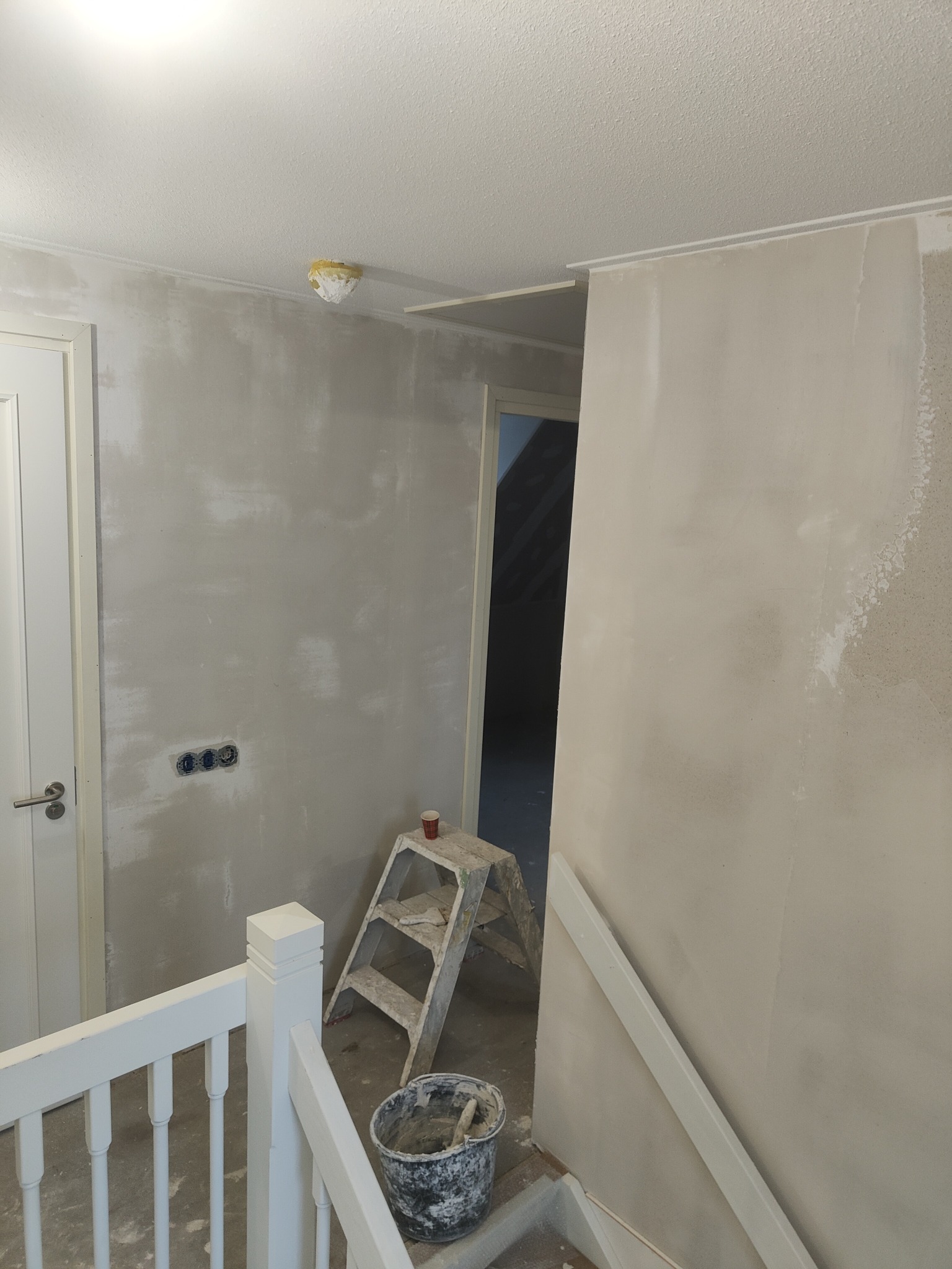 Ściana w trakcie szpachlowania z widocznymi śladami po nałożonym gipsie, drabina malarska, wiadro z zaprawą, zabezpieczone gniazdka elektryczne i fragment białej balustrady.