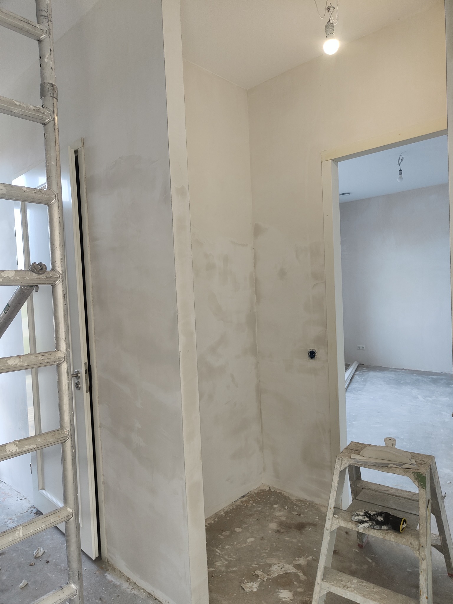 Wnętrze w trakcie szpachlowania: ściany pokryte szpachlą, aluminiowa drabina z lewej strony i mały stołek z narzędziami i rękawicami z prawej strony, surowa żarówka na suficie.