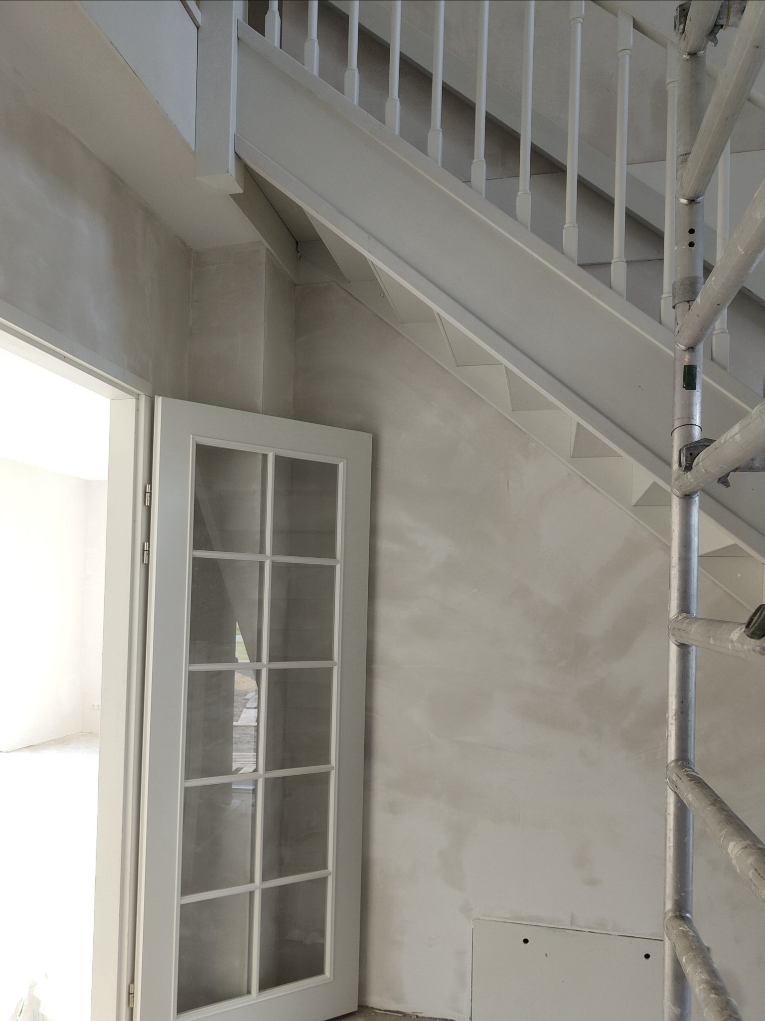 Wnętrze w trakcie prac wykończeniowych: ściana po szpachlowaniu, widoczne rusztowanie, białe schody z balustradą i częściowo otwarte białe drzwi.