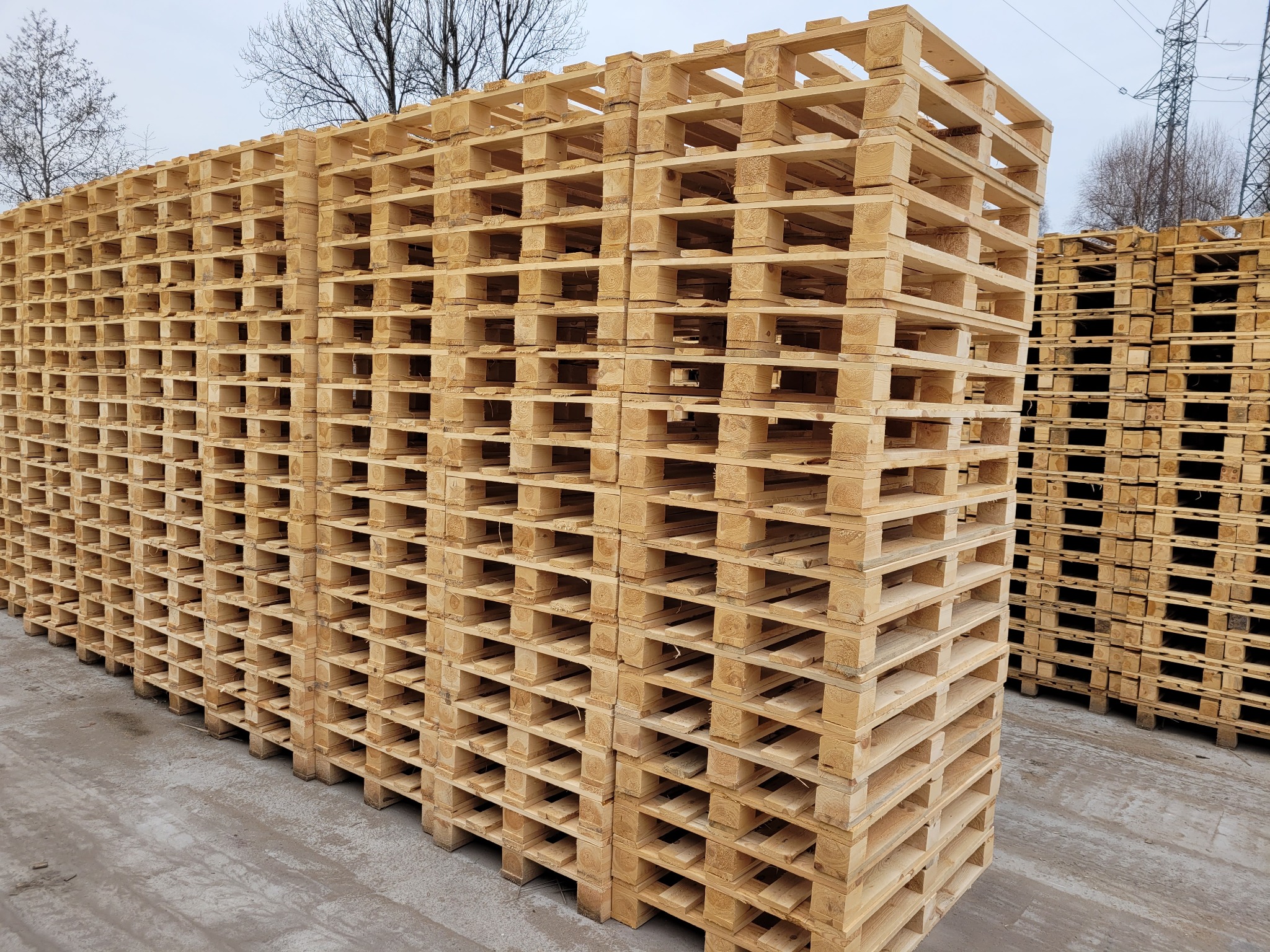 Stosy jasnych, drewnianych palet ułożonych na zewnątrz, z widocznymi słupami wysokiego napięcia w tle.