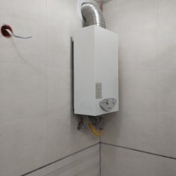 LOUIS BLUES PAVEL BAROUSKI - Biały gazowy podgrzewacz wody zamontowany na ścianie w łazience wyłożonej beżowymi płytkami, z widoczną rurą odprowadzającą spaliny i żółtym przewodem gazowym.