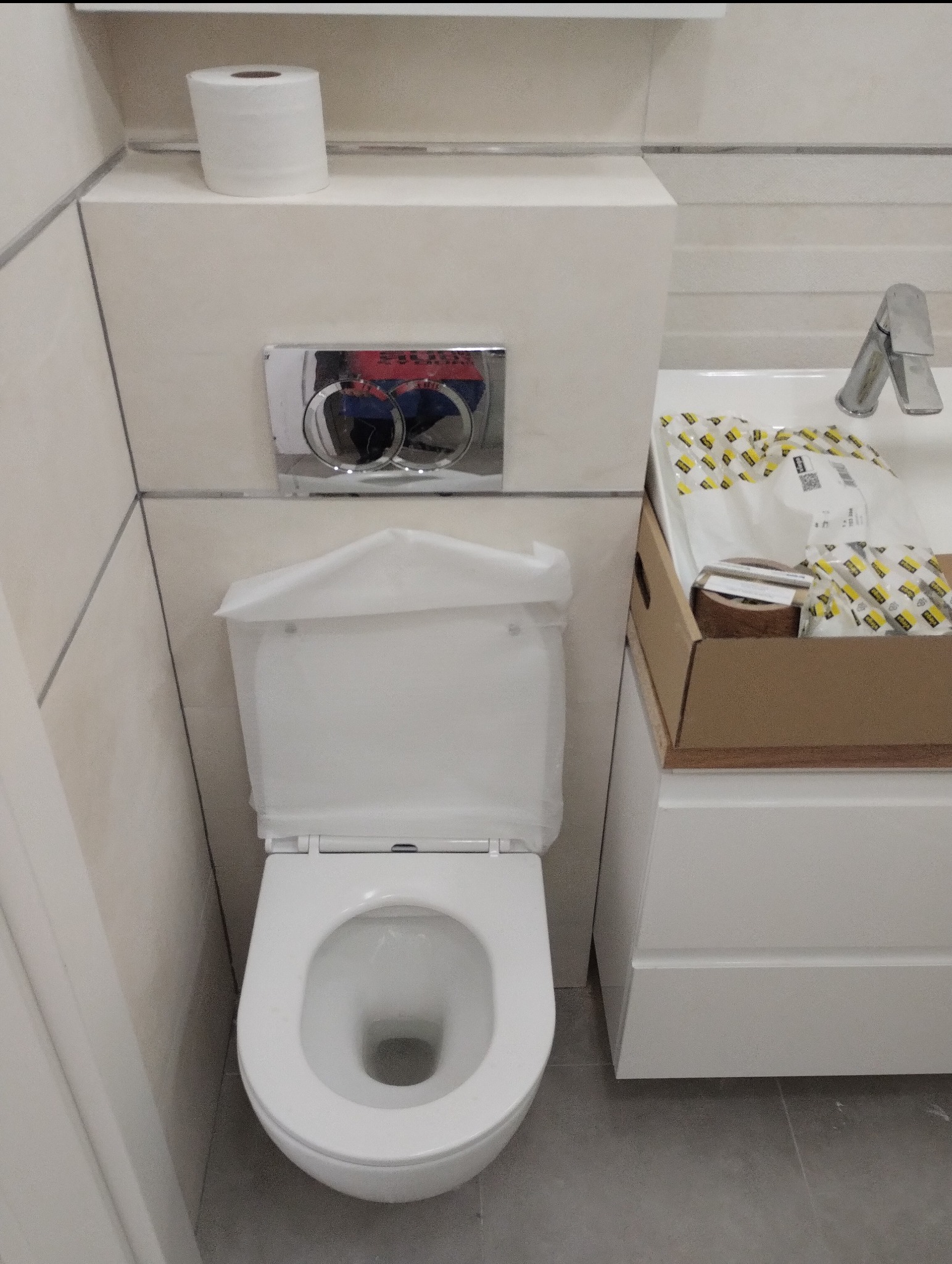 Nowoczesna toaleta podwieszana z chromowanym przyciskiem spłukującym, rolka papieru toaletowego na obudowie, umywalka z baterią i akcesoriami w kartonie, całość w jasnej łazience po remoncie.