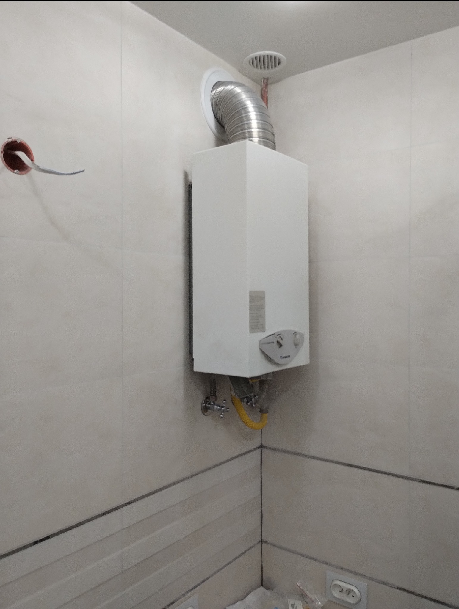 Biały gazowy podgrzewacz wody zamontowany na ścianie w łazience wyłożonej beżowymi płytkami, z widoczną rurą odprowadzającą spaliny i żółtym przewodem gazowym.