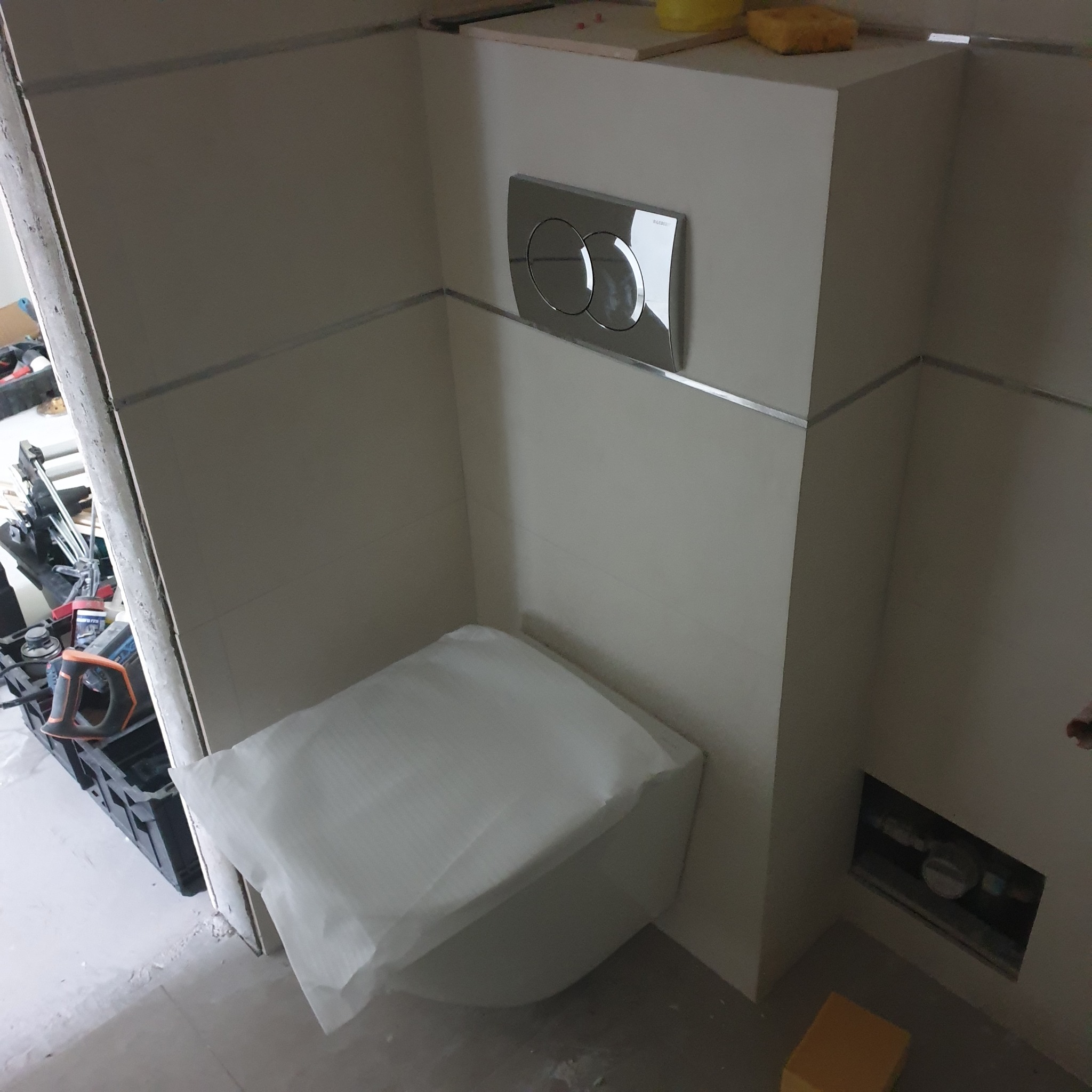 Wisząca toaleta w trakcie montażu, zabezpieczona folią, z widocznym przyciskiem spłukującym Geberit i narzędziami w tle.