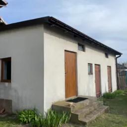 Jasnoszary budynek gospodarczy z brązowymi rynnami, oknami i drzwiami; widoczne schody wejściowe i otwarte drzwi samochodu dostawczego w tle.