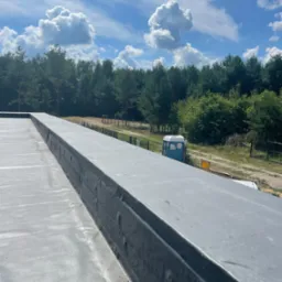 Szary, płaski dach pokryty membraną bitumiczną, widok z góry na las i błękitne niebo z białymi chmurami, w tle przenośna toaleta.