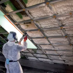Pracownik w kombinezonie ochronnym aplikuje białą pianę izolacyjną na poddasze z widocznymi metalowymi profilami i oknem dachowym.