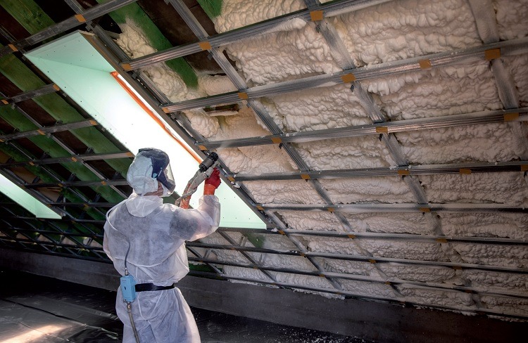 Pracownik w kombinezonie ochronnym aplikuje białą pianę izolacyjną na poddasze z widocznymi metalowymi profilami i oknem dachowym.