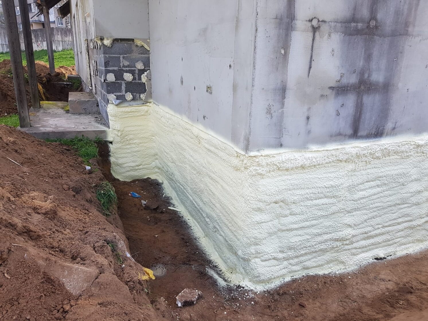 Izolacja fundamentów domu jednorodzinnego, pokryta warstwą pianki poliuretanowej, widoczna wykopana ziemia wokół budynku.