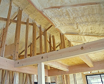 Poddasze domu w trakcie ocieplania, widoczne drewniane krokwie i biała pianka izolacyjna pokrywająca sufit i ściany.
