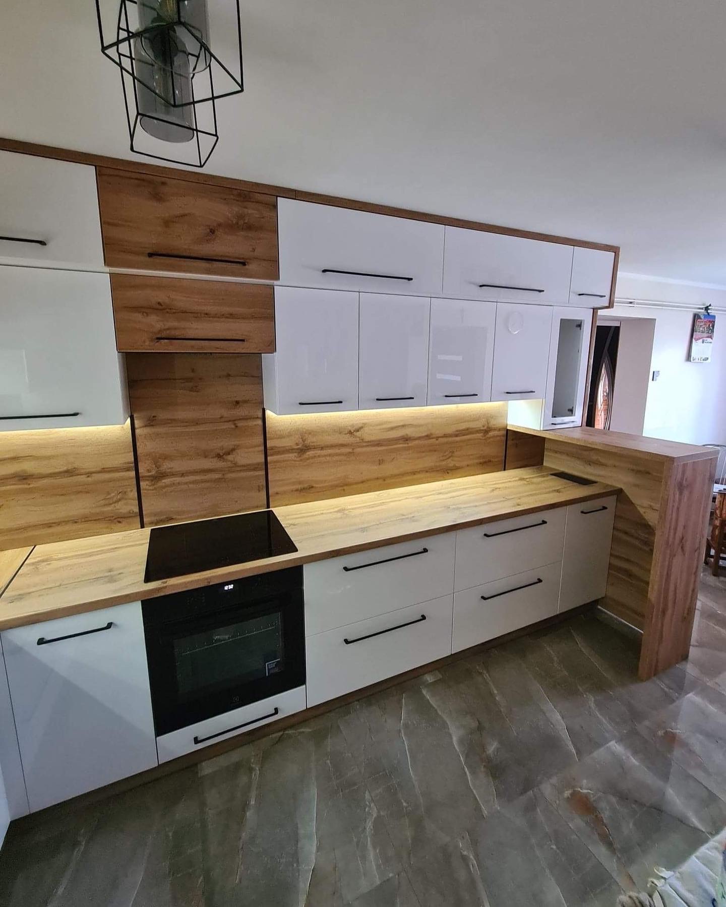 Nowoczesna kuchnia z białymi szafkami i drewnianymi akcentami, czarnymi uchwytami, oświetleniem LED pod szafkami wiszącymi i płytą indukcyjną, widok na marmurową podłogę.