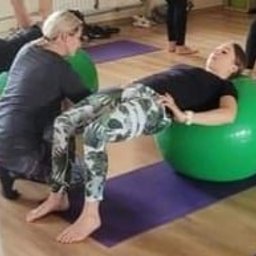 PILATES Reha MAGDALENA STAWARZ - Kobieta ćwiczy unoszenie bioder na zielonej piłce rehabilitacyjnej pod nadzorem instruktorki, w tle grupa osób wykonuje podobne ćwiczenia na piłkach.