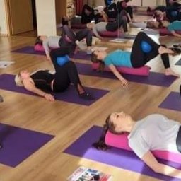 PILATES Reha MAGDALENA STAWARZ - Grupowe zajęcia jogi z wykorzystaniem wałków i piłek, uczestniczki w czarnych legginsach i różnokolorowych koszulkach ćwiczą na fioletowych matach w jasnej sali.