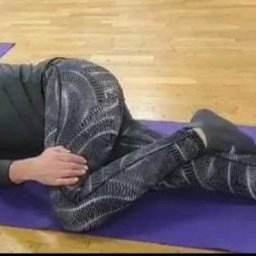 PILATES Reha MAGDALENA STAWARZ - Kobieta w czarnym ubraniu leżąca na fioletowej macie, wykonująca pozycję skrętu kręgosłupa podczas zajęć w grupie.