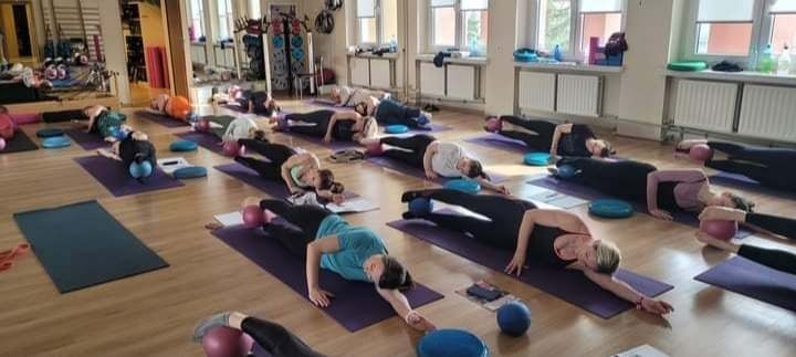 Grupa osób ćwiczy na matach w przestronnym, jasnym pomieszczeniu; kobiety leżą na boku, opierając się na jednym ramieniu, z piłkami pod biodrami, podczas zajęć grupowych.