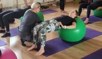 Kobieta ćwiczy unoszenie bioder na zielonej piłce rehabilitacyjnej pod nadzorem instruktorki, w tle grupa osób wykonuje podobne ćwiczenia na piłkach.