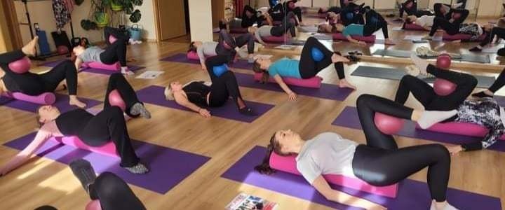 Grupowe zajęcia jogi z wykorzystaniem wałków i piłek, uczestniczki w czarnych legginsach i różnokolorowych koszulkach ćwiczą na fioletowych matach w jasnej sali.