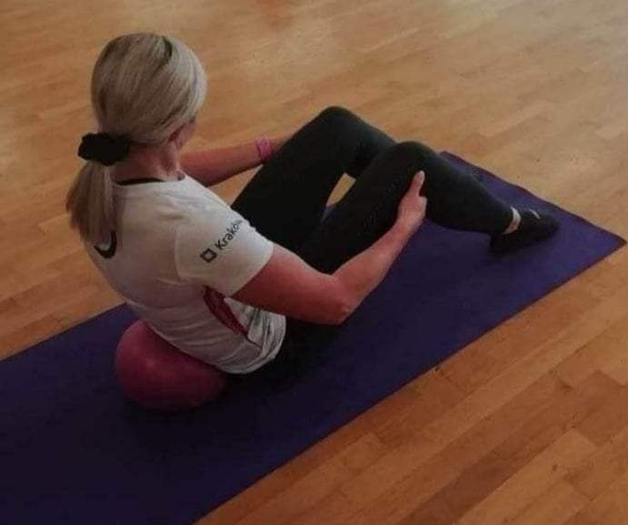 Kobieta siedzi na różowej piłce gimnastycznej na fioletowej macie, wykonując ćwiczenie wzmacniające mięśnie brzucha, widok z tyłu, na drewnianej podłodze.