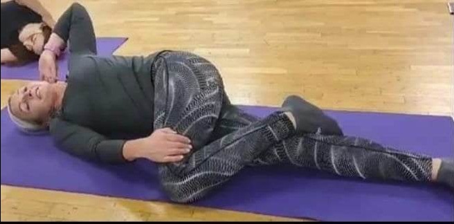 Kobieta w czarnym ubraniu leżąca na fioletowej macie, wykonująca pozycję skrętu kręgosłupa podczas zajęć w grupie.