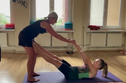 Kobieta pomaga drugiej osobie w rozciąganiu mięśni nóg podczas ćwiczenia na macie w jasnym pomieszczeniu z oknami.