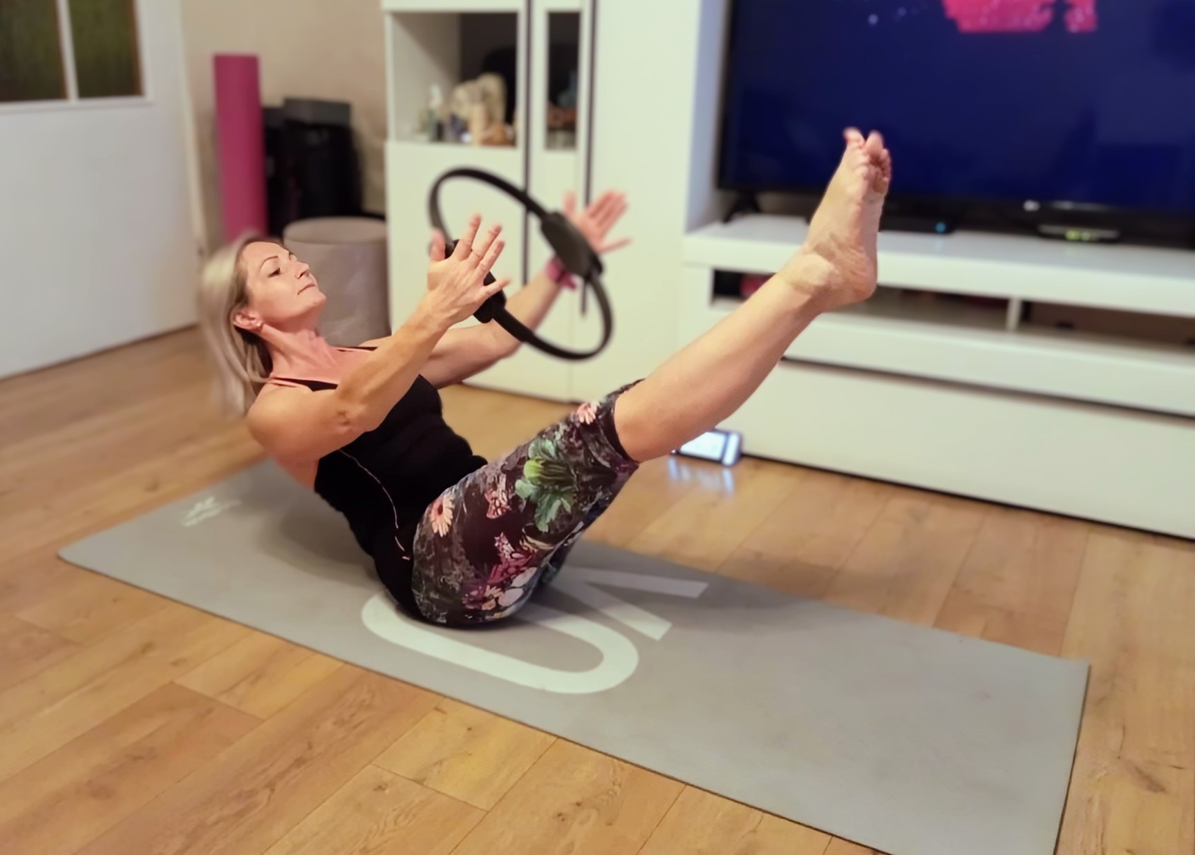 Kobieta w pozycji V-up na macie, ćwiczy z pierścieniem do pilatesu. W tle meble i telewizor. Ćwiczenia w domu.